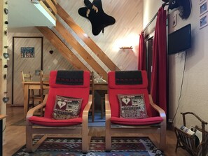 Ein gemütliches Zimmer mit schrägverdeckter Decke, zwei rote Polsterstühle mit gemusterten Kissen, ein Holztisch und ein Fernseher an der Wand.