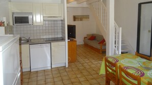 Una cocina compacta con armarios blancos, un microondas y un lavavajillas. Hay una zona de comedor con una mesa y sillas, y una escalera que conduce a un nivel superior.