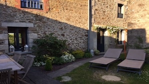Un bâtiment en pierre doté d’un patio comprenant une table et des chaises, un petit jardin et deux chaises longues.