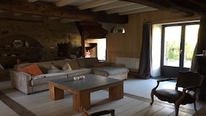 Un salon rustique avec un canapé en bois, une table basse et un fauteuil. On y trouve une cheminée et une grande fenêtre avec des rideaux.