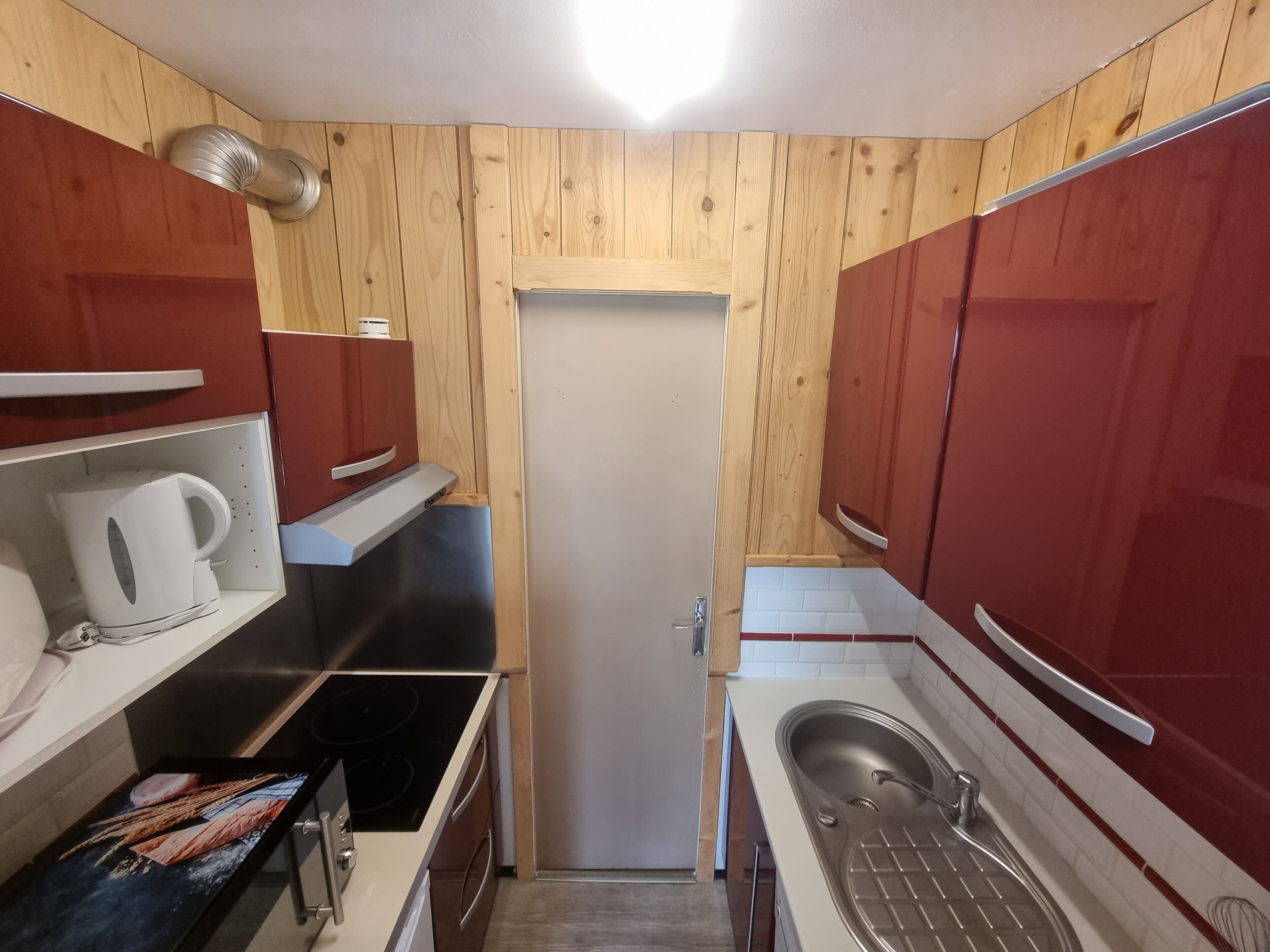 Pieni, käytännöllinen keittiö, jonka seinät ovat puuta, ovi on valkoinen ja kaapit punaisia.