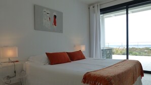A bedroom with a large bed, two red pillows, a white blanket, a painting on the wall, and a view of the sea through a large window. -  MODERN VILLA 10 PERONNES SWIMMING POOL 10MN FROM THE BEACH AND SPERONE GOLF VIEW  (Bonifacio)