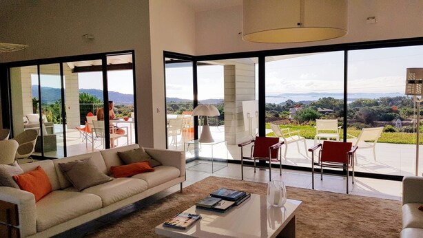 A spacious living room with a large window offering a view of the outdoors, a sofa with cushions, a coffee table, and a dining area with chairs. -  MODERN VILLA 10 PERONNES SWIMMING POOL 10MN FROM THE BEACH AND SPERONE GOLF VIEW  (Bonifacio)