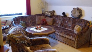 A living room with a large, patterned sofa, a matching armchair, and a small round coffee table with a lace doily.