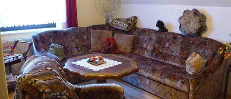 A living room with a large, patterned sofa, a matching armchair, and a small round coffee table with a lace doily.