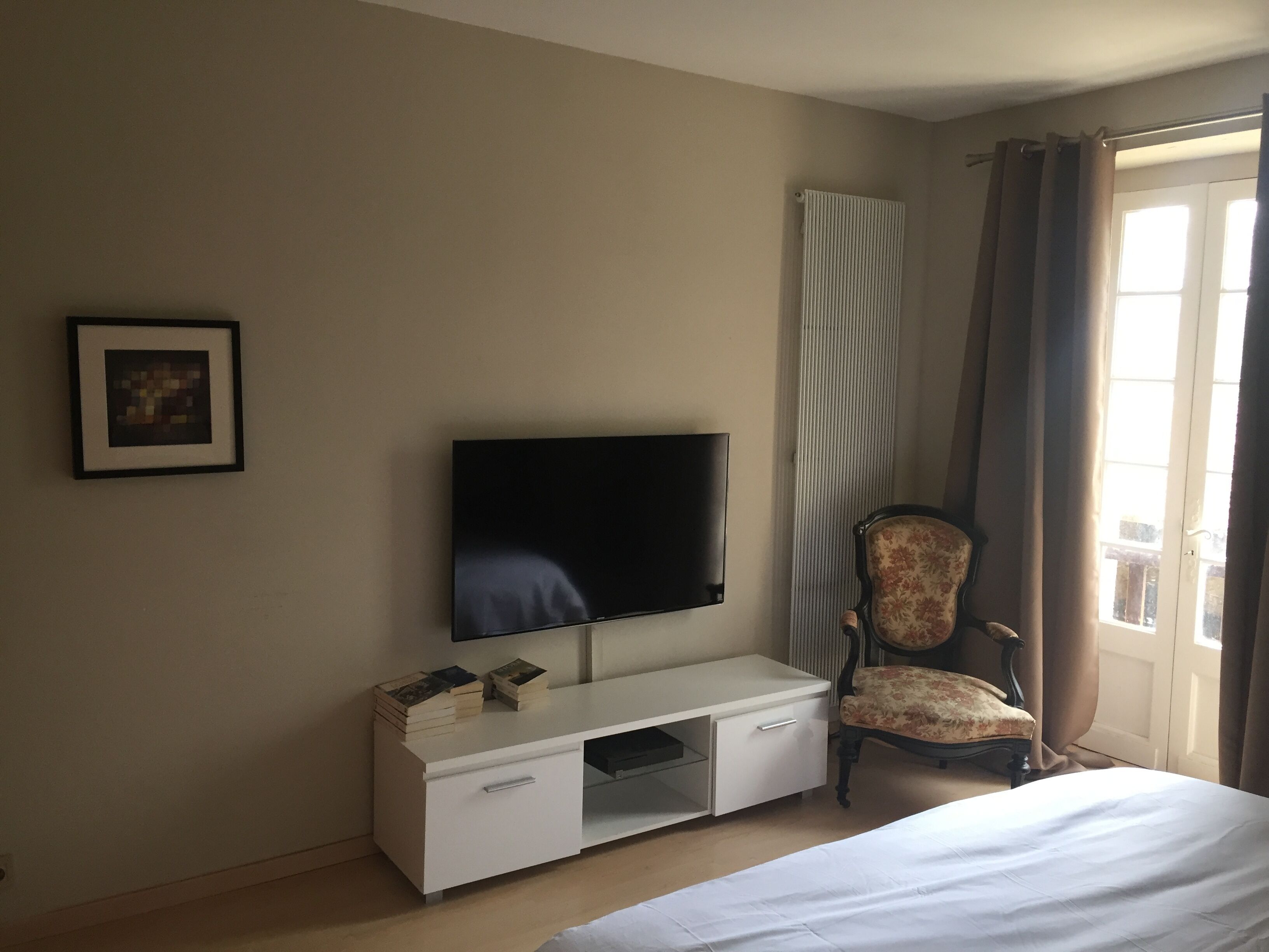 Une chambre d’hôtel avec un lit, un téléviseur posé sur une commode blanche, une chaise et une fenêtre avec des rideaux.
