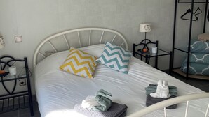 Un lit bien fait, avec une literie blanche, deux coussins décoratifs, un luminaire fixé au mur et une petite étagère sur laquelle reposent une bougie et un livre.