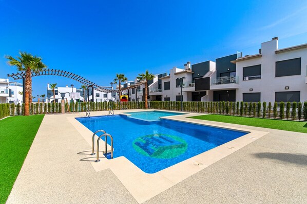 A modern pool area with a clear blue pool, a palm tree, and a white building with balconies.