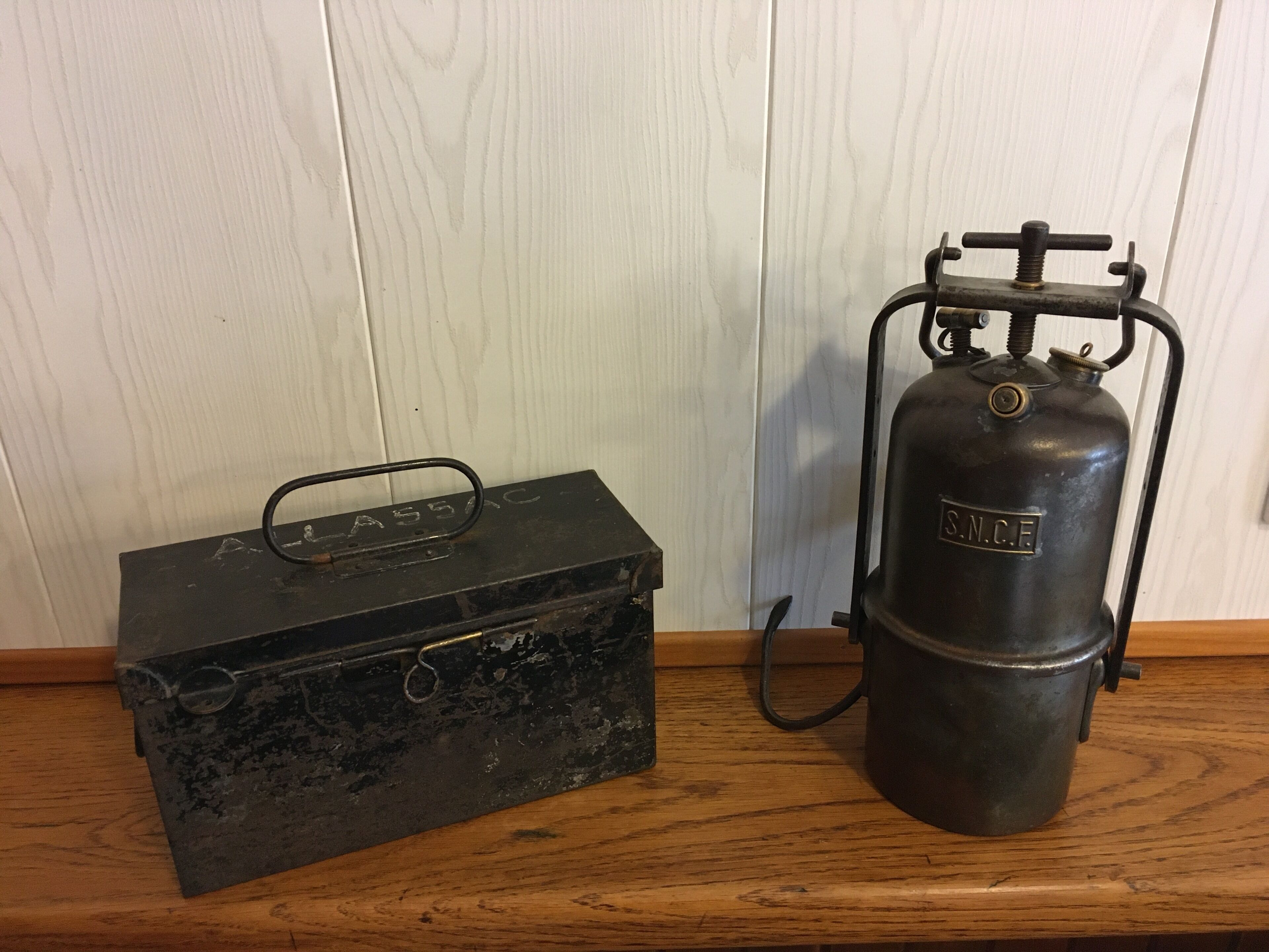A vintage black metal box with a handle and a metal gas cylinder with a valve and hose.