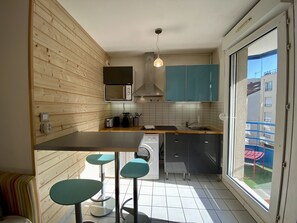 A compact kitchen with wooden paneling, a microwave, a toaster, a sink, and a washing machine. There are three teal stools and a window with a view of a balcony.