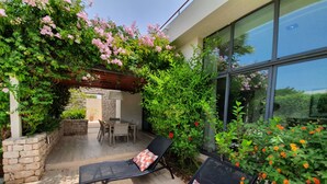 Un patio con dos sillones de descanso y una mesa, rodeado de abundante vegetación y plantas con flores.