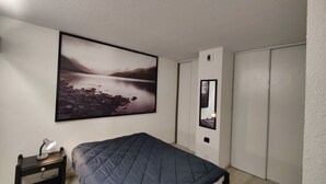 Ein Schlafzimmer mit einem Bett, einem Nachttisch, einer Schreibtischlampe, einem großen gerahmten Landschaftsbild, einem Spiegel und einem Kleiderschrank.