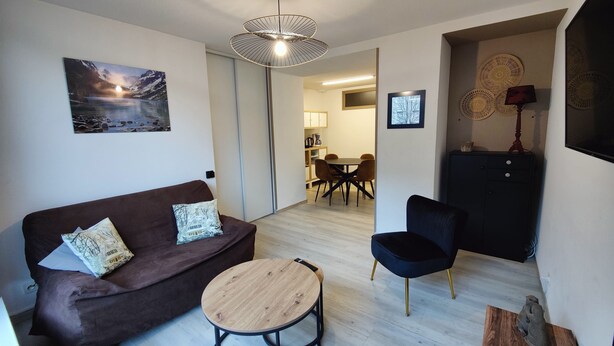 Ein modernes Wohnzimmer mit einem braunen Sofa, einem runden Couchtisch und einem schwarzen Sessel. Im Hintergrund ist ein Essbereich mit Tisch und Stühlen erkennbar.