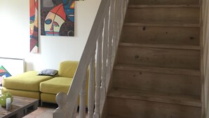 Una escalera con escalones de madera y barandillas blancas que conduce a un piso superior. Un sofá amarillo y una pequeña mesa con un jarrón verde son visibles en primer plano.