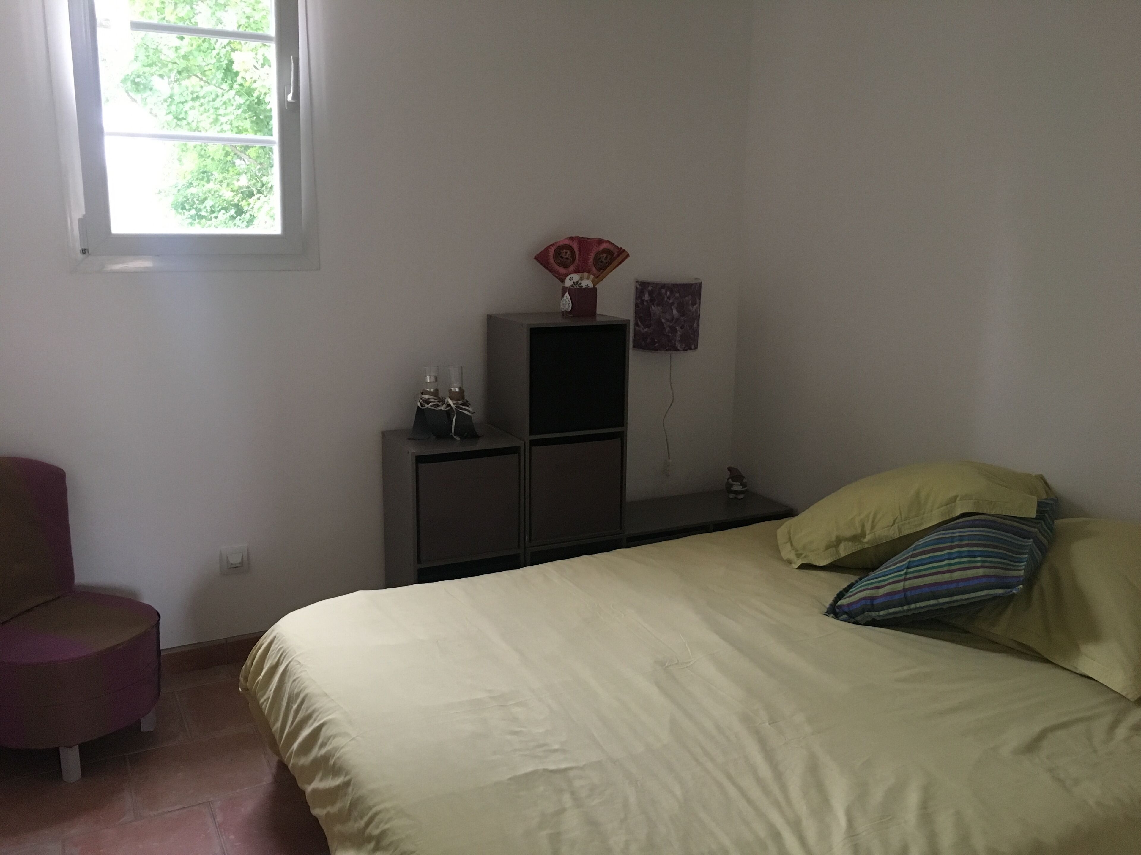 A bedroom with a bed, a chair, a small cabinet, and a window.