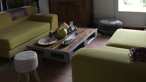 Un salón con un sofá amarillo, una mesa de centro de madera con objetos decorativos, un televisor sobre un mueble de madera y un ventanal con vistas al verdor.