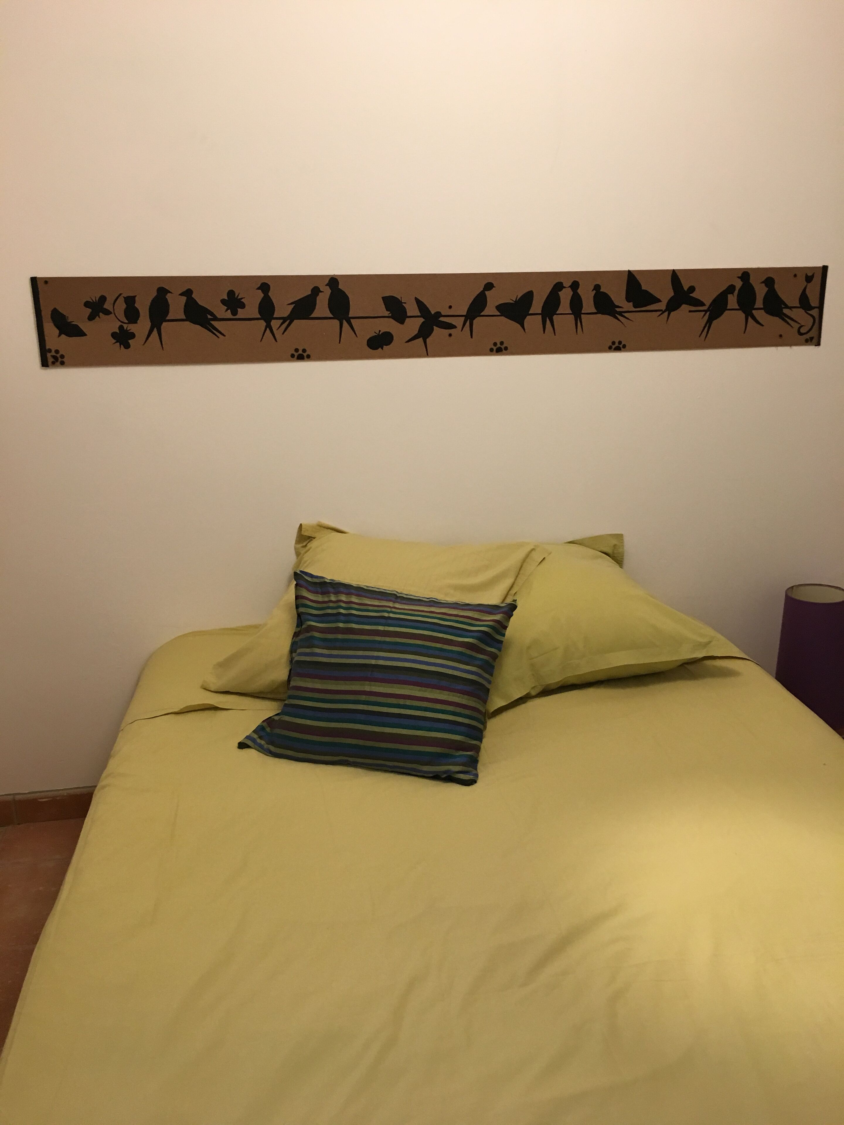 A bed with yellow bedding and a striped pillow, a wall decoration with bird silhouettes, and a purple cylindrical object on the right.