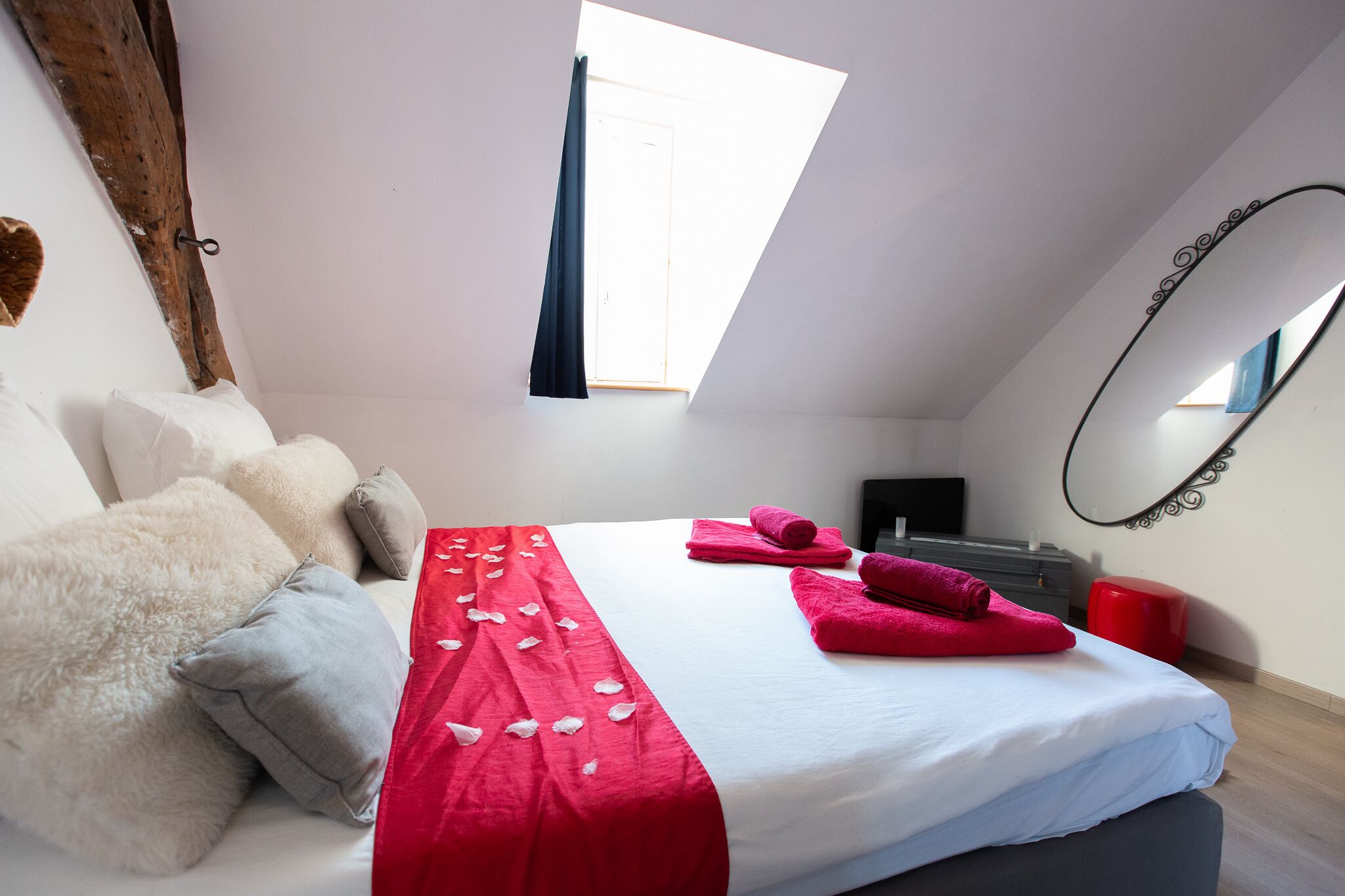 A bedroom with a bed, pillows, a red and white bedspread, a red stool, a mirror, and a window.