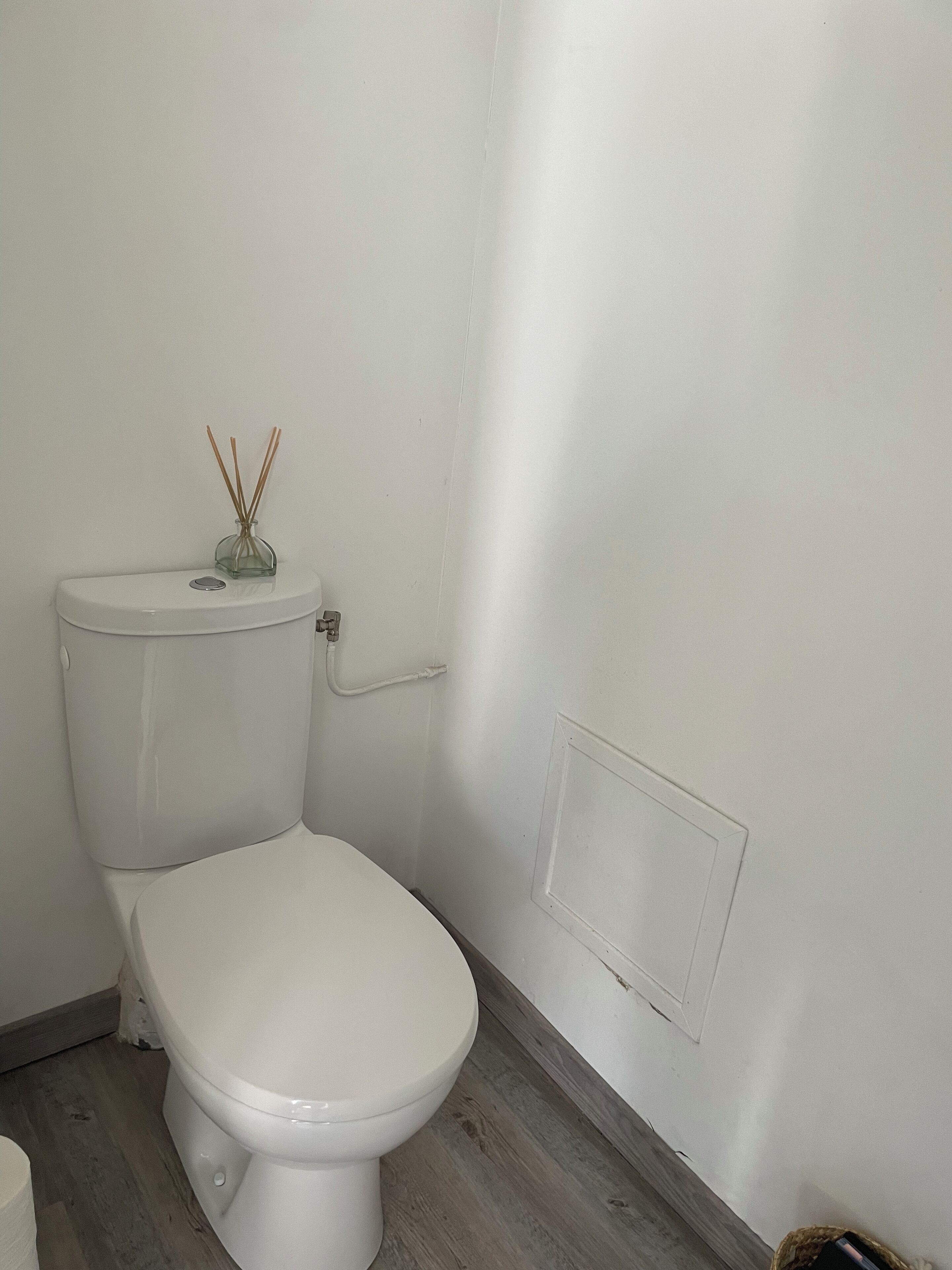 Des toilettes blanches, avec un petit récipient en verre contenant des diffuseurs à bâtonnets posé sur le réservoir, un mur blanc et un plancher en bois.