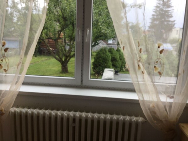 A window with sheer curtains and a view of a green landscape with trees and a house.