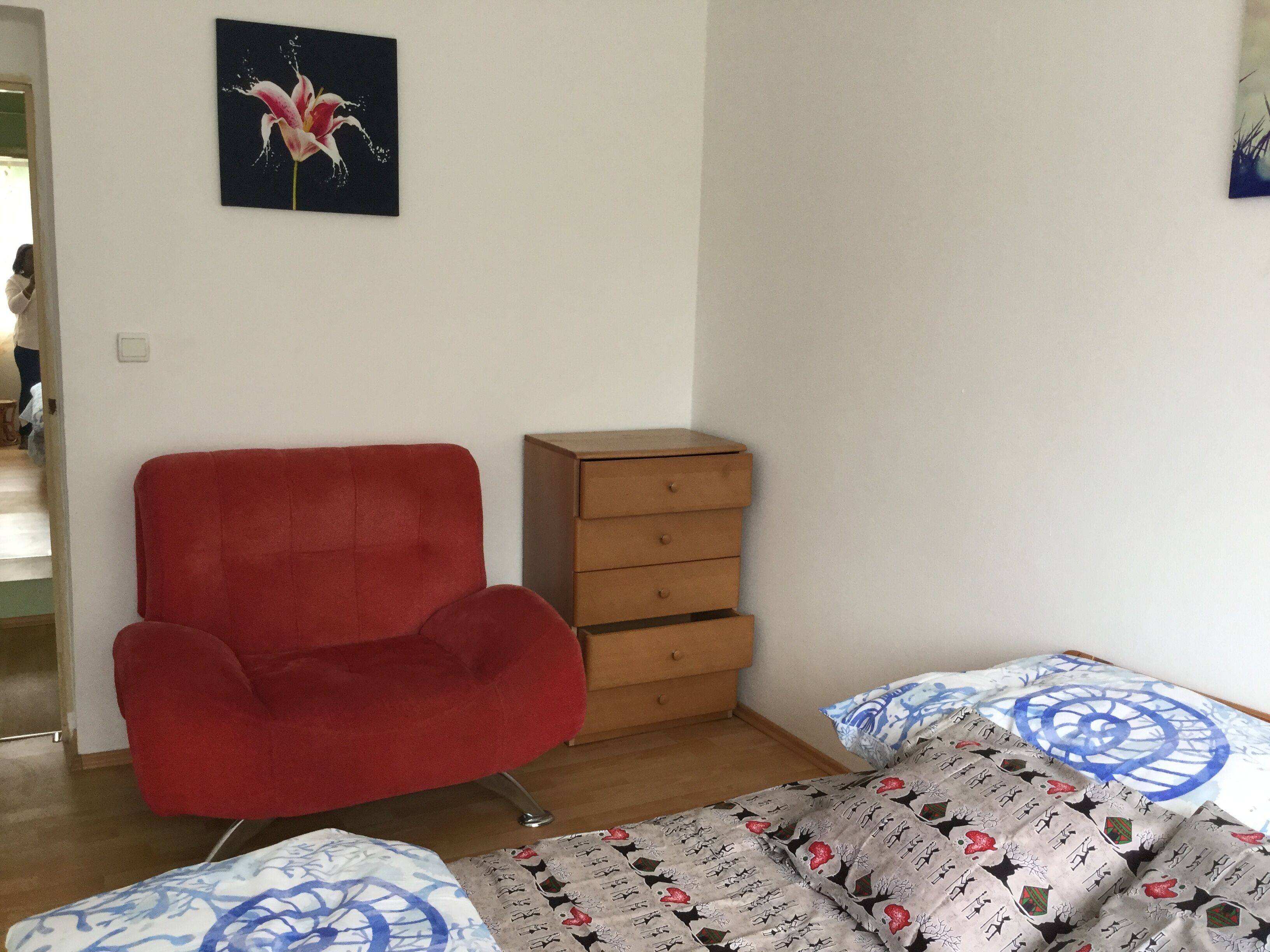 A room with a red armchair, a wooden chest of drawers, and a bed with patterned bedding.