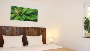 A bedroom with a wooden headboard, two white pillows, a bed with a beige bedspread, a wall-mounted picture of green leaves, a bedside lamp, and a window with a plant. - Modern apartment with terrace and use of sauna (Burgbernheim)