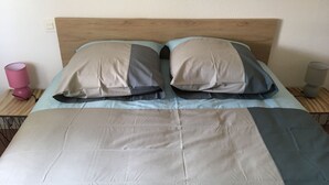 Een bed met een houten hoofdbord, twee kussens, een lichtblauwe lakenset en een grijze deken. Er zijn twee nachtkastjes, waarvan er eentje een roze lamp heeft en de ander een grijze.