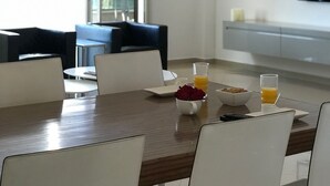 A modern dining area with a wooden table, white chairs, and a chandelier. There is a bowl of fruit and a glass of orange juice on the table. - Véronique luxuriou's penthouse (Eilat)