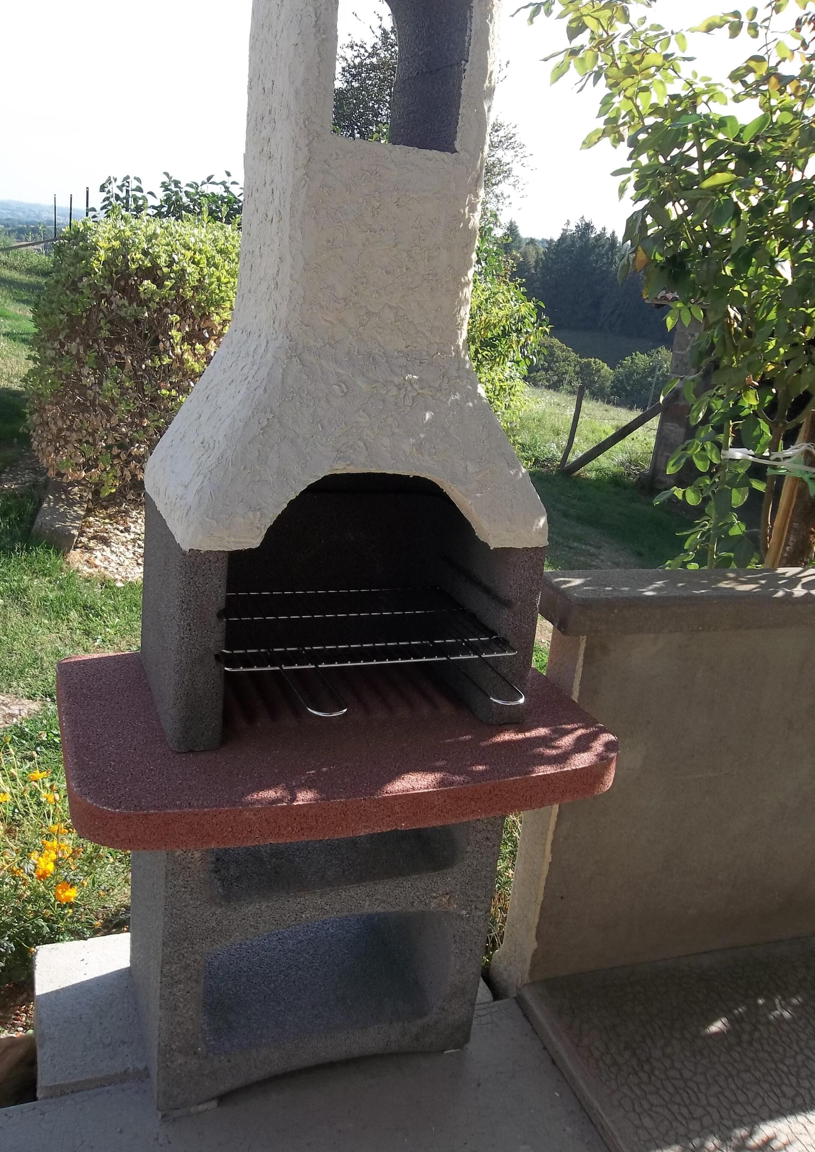 A stone-built outdoor oven with a metal rack, set on a concrete base, surrounded by greenery.