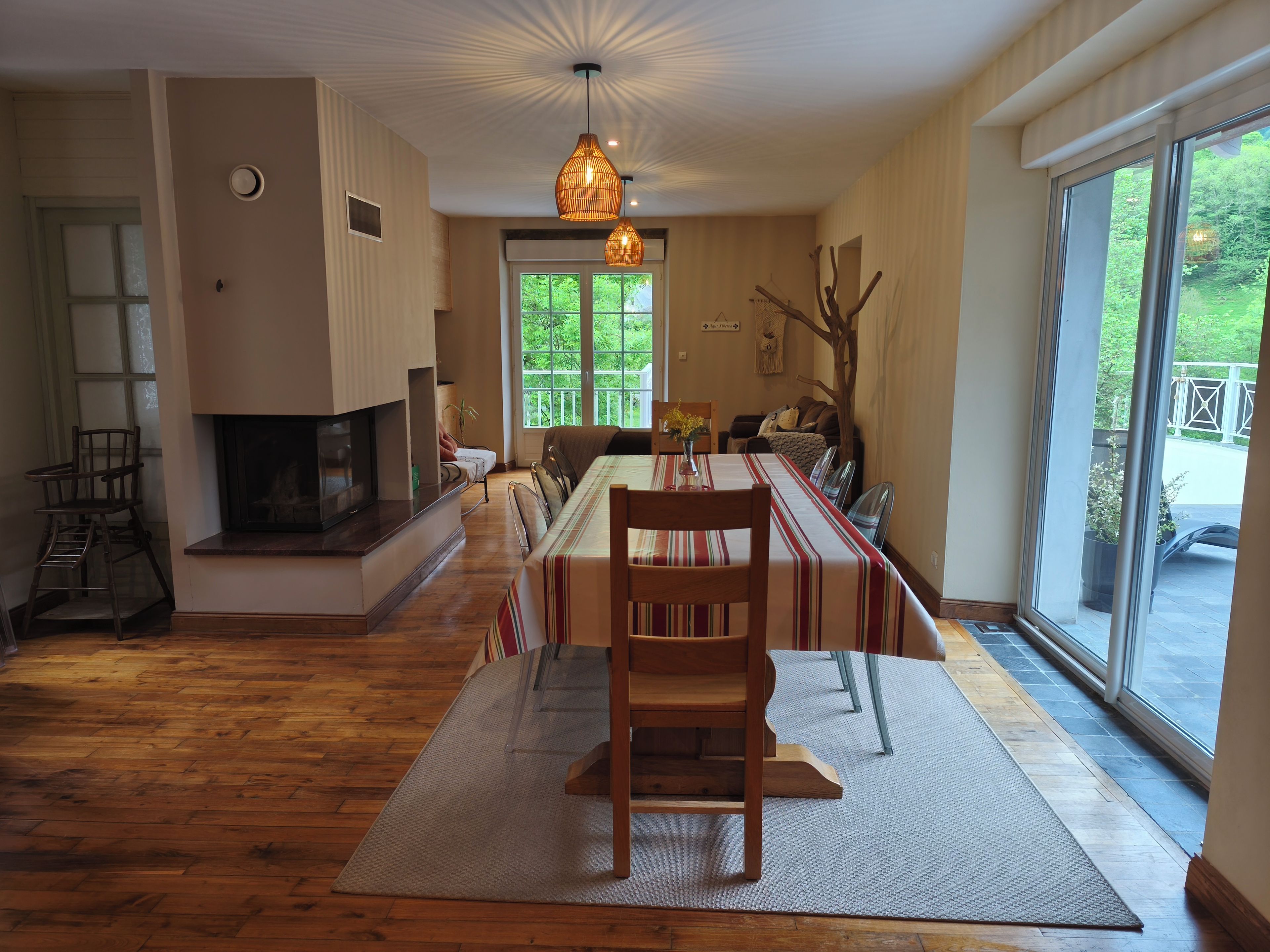 A dining area with a long table, chairs, a fireplace, and a view through a large window.