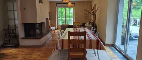A dining area with a long table, chairs, a fireplace, and a view through a large window.