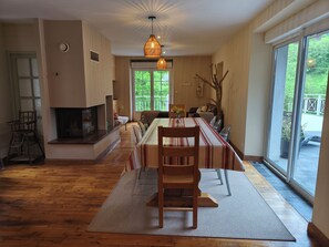 A dining area with a long table, chairs, a fireplace, and a view through a large window.