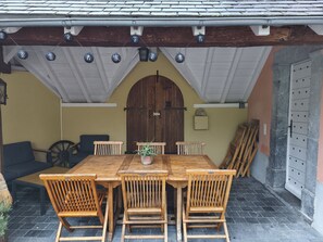 A wooden table with chairs, a potted plant, and a large wooden door in a covered outdoor area.