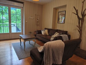 Een woonkamer met een bruine bank, een houten salontafel, een vloerkleed met patroon en een groot raam met uitzicht op groen.
