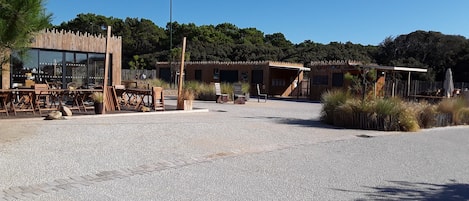 A spacious outdoor area with a gravel ground, wooden furniture, and a building with large windows and a wooden fence.
