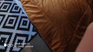 A neatly made bed with a brown quilt and two rolled towels, a patterned rug, a wall-mounted shelf with books, and a door with heart-shaped cutouts.