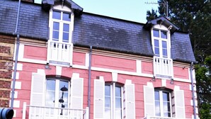 A two-story building with a red facade and white trim, featuring multiple windows with shutters, a balcony, and a chimney.