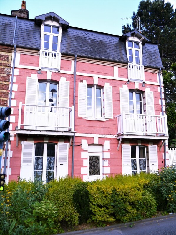 A two-story building with a red facade and white trim, featuring multiple windows with shutters, a balcony, and a chimney.