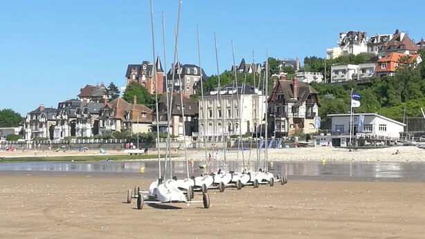 A beach with several sailboats docked on the sand, a row of houses in the background, and a clear blue sky.