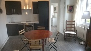 A compact kitchen with dark cabinets, a stainless steel range hood, a built-in oven, and a small dining area with a wooden table and chairs. - Charming cottage 200 meters from the beach, in beautiful wooded property (Veulettes-sur-Mer)