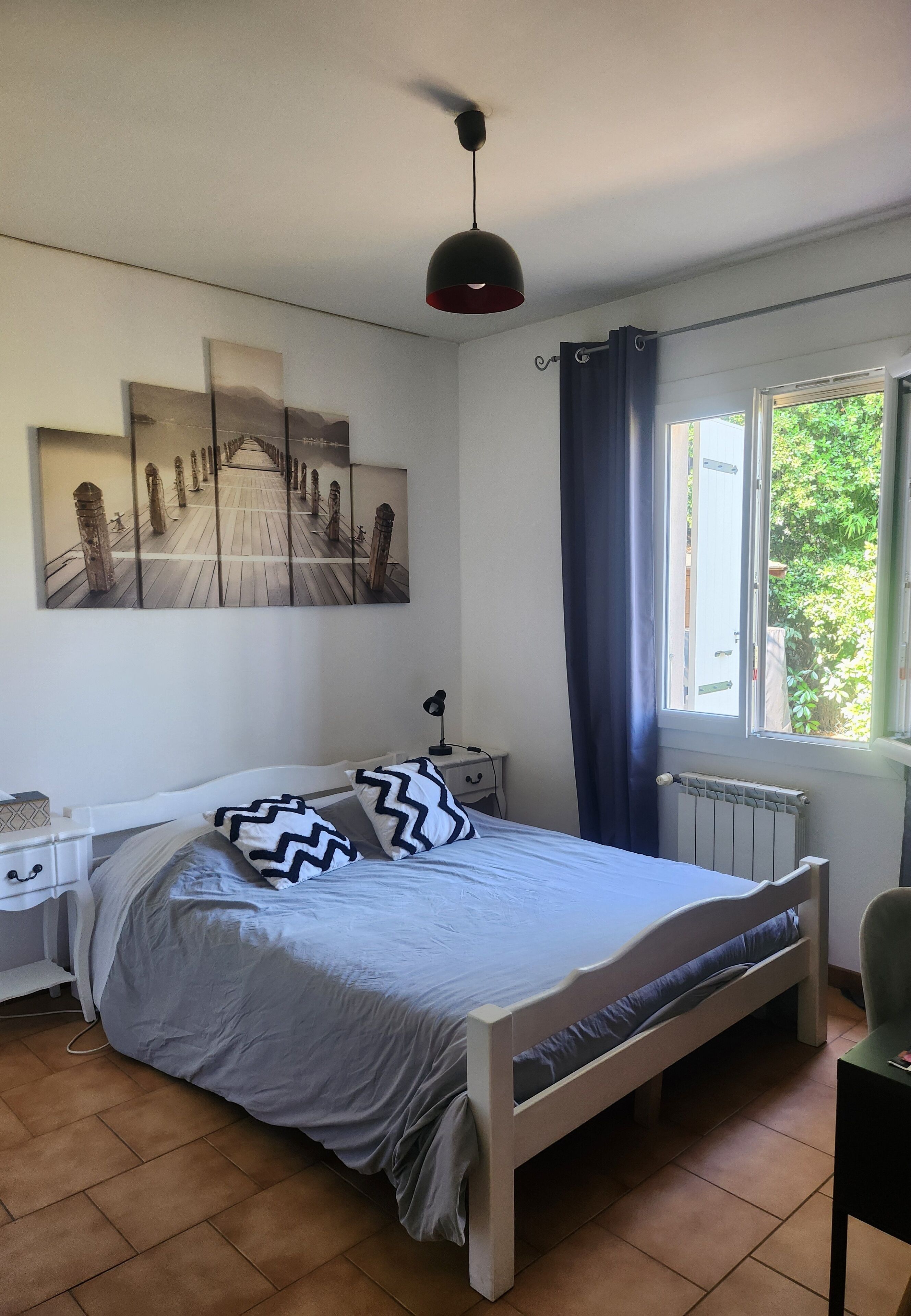 A bedroom with a bed, a nightstand, a lamp, a window with curtains, and a radiator.