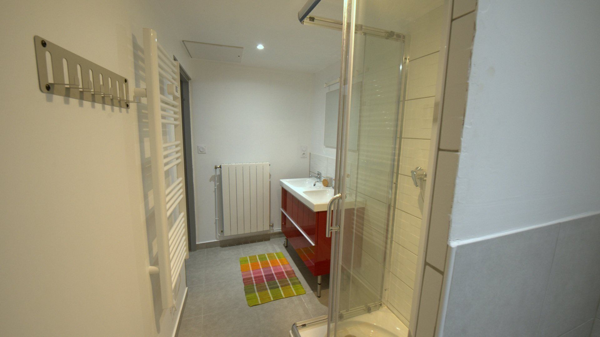 Un baño estrecho con cabina de ducha de vidrio, un lavamanos blanco con espejo y un radiador en la pared.