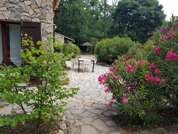 A stone patio with a table and chairs, surrounded by lush greenery and flowering plants.