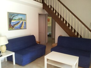 A living room with two blue sofas, a white coffee table, and a painting of a field. A staircase with wooden railings is visible in the background.