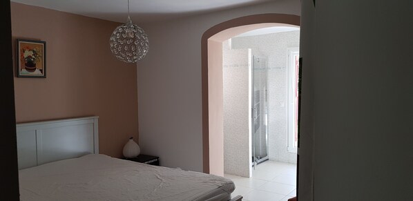 A bedroom with a bed, a nightstand, a decorative lamp, a framed picture on the wall, and a shower area visible through an archway.