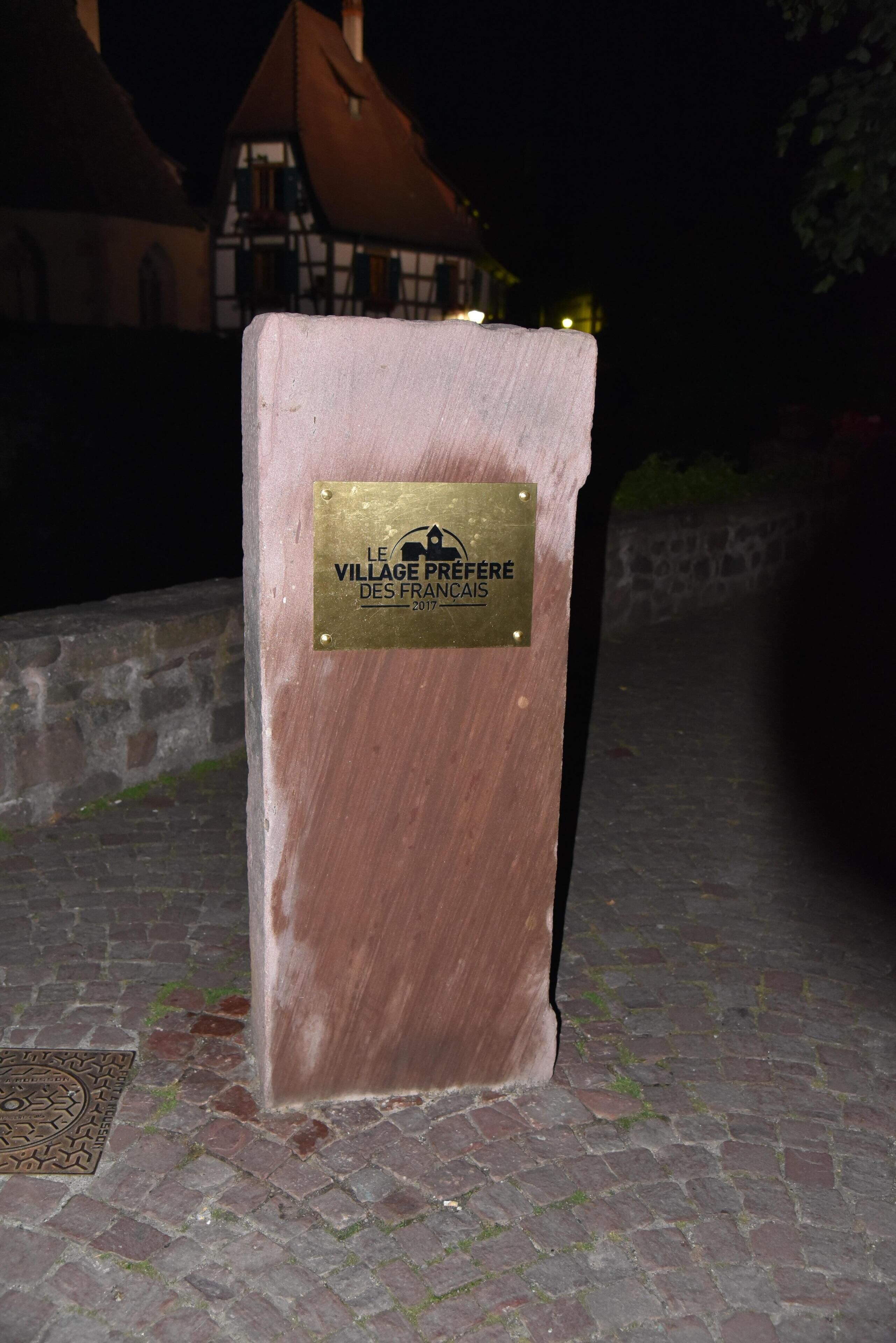 Une plaque fixée à une structure en pierre, avec un bâtiment ancien en arrière-plan.
