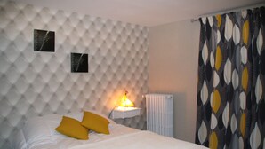 A bedroom with a tufted headboard, a bedside table with a lamp, a bed with white bedding and two yellow pillows, and a patterned curtain. - House completely renovated in the countryside in tourist village (Saint-Evroult-Notre-Dame-du-Bois)