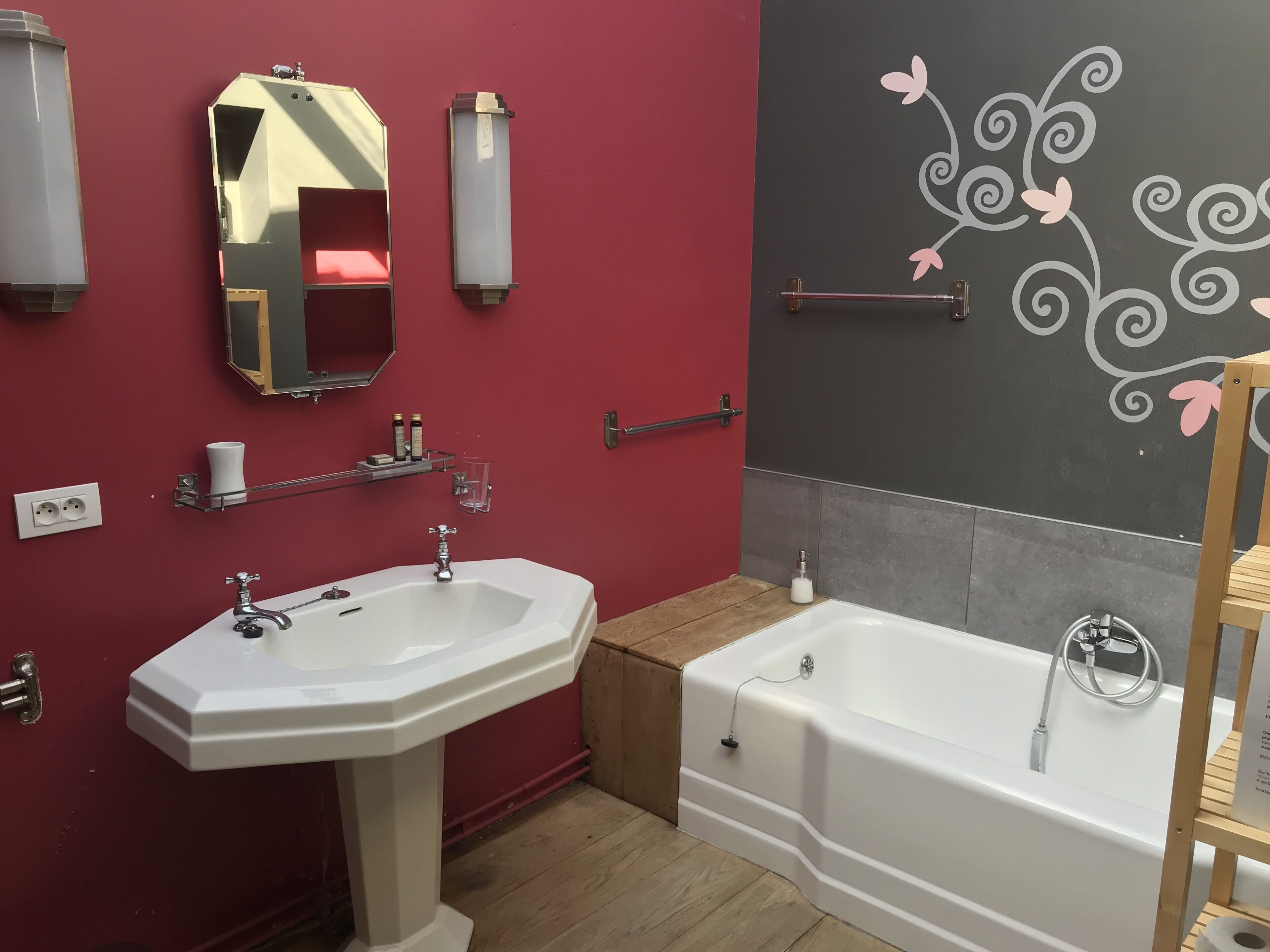 Een badkamer met een wastafel op een voetstuk, een badkuip, een spiegel en een decoratieve wand met bloemmotieven.
