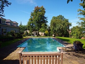 Una piscina rectangular con muelle de madera en primer plano, rodeada de césped cuidado y árboles.