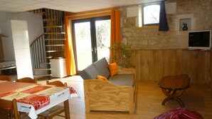 Una sala de estar acogedora con un sofá de madera, una mesa de comedor con mantel rojo, una zona de cocina con estufa y una escalera que sube a otro nivel.